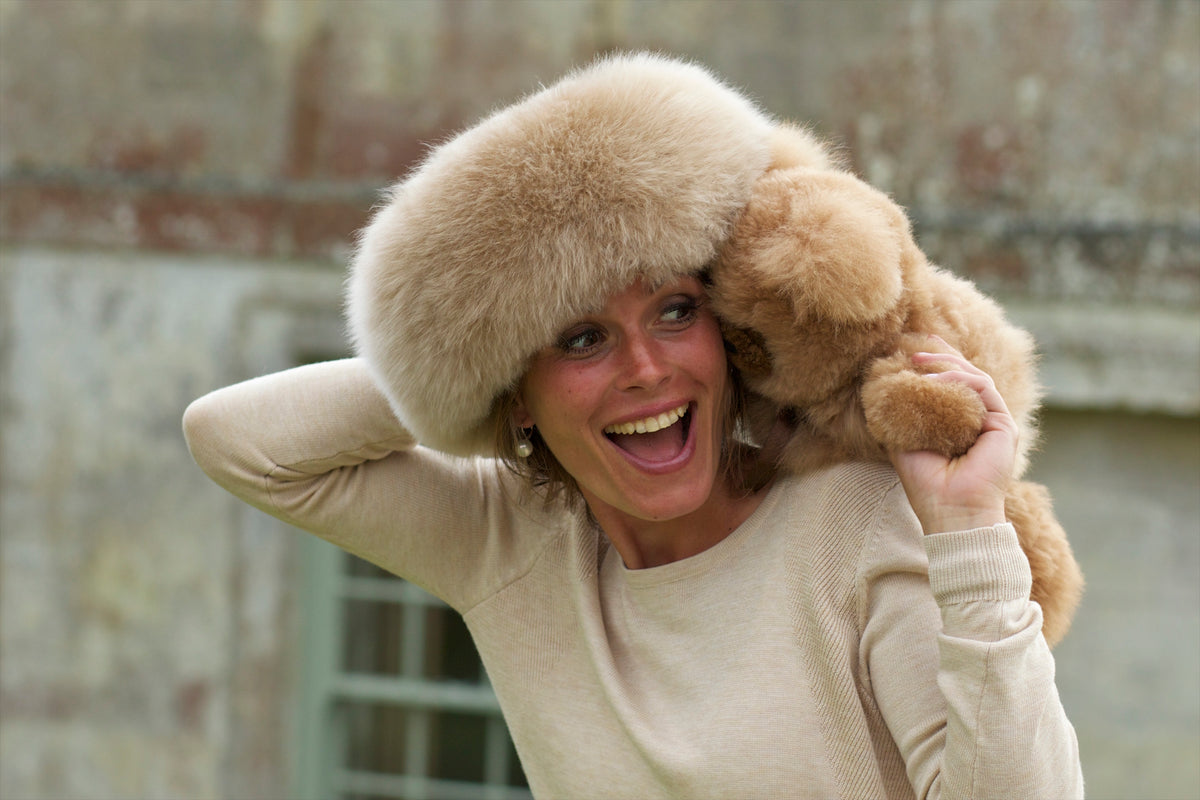 A woman wearing a large alpaca fur hat and cream sweater holds a tan alpaca fur teddy bear against her shoulder.